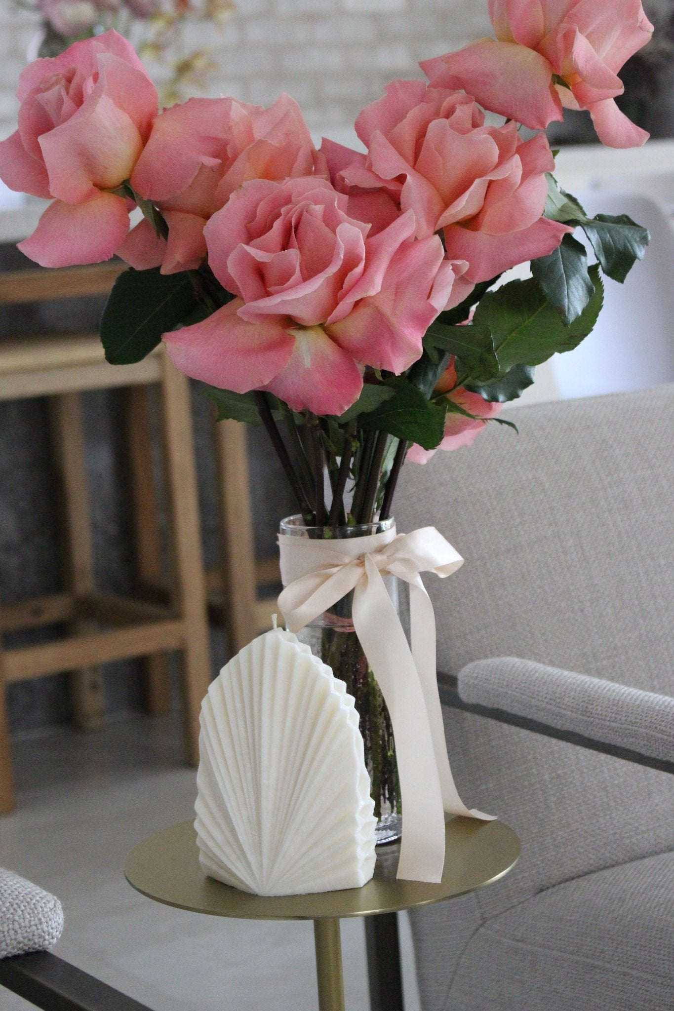 Palm leaf shaped candle Aria in Ivory (white) colour displayed on side table near a vase of pink roses.