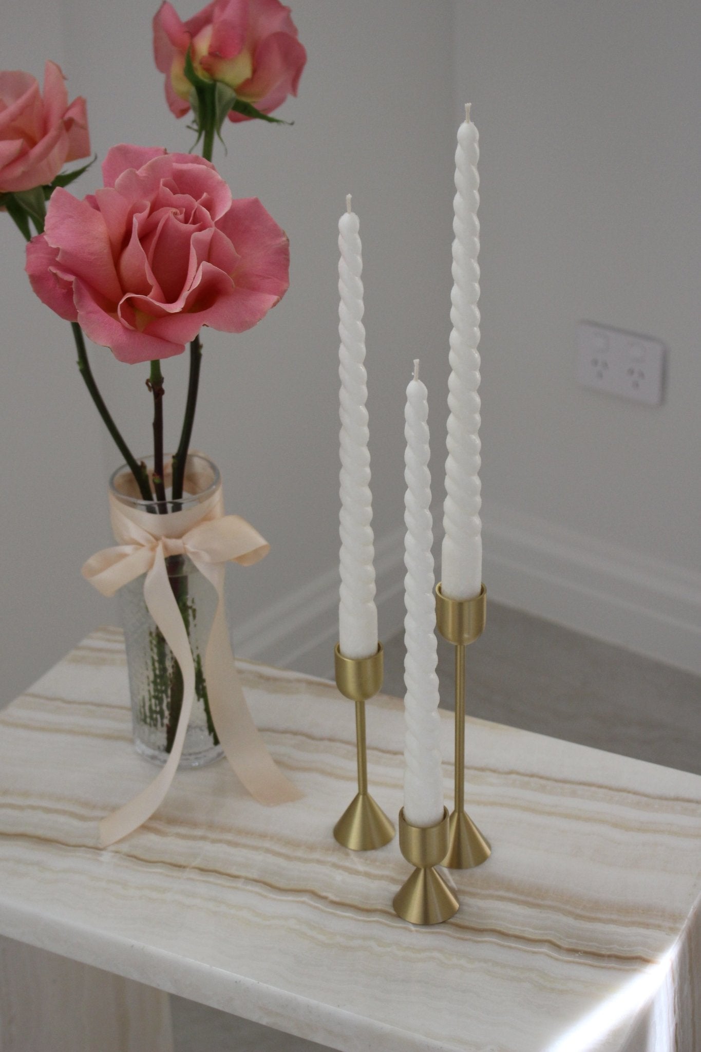 Twisty Ariel taper candle in Ivory (white) colour displayed in a trio on brass candlestick holders set (also known as Lumiere candle holder set). They are positioned on a marble side table near a vase of roses.