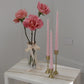Twisty Ariel taper candle in Peony (pastel pink) colour displayed in a trio on brass candlestick holders set (also known as Lumiere candle holder set). They are positioned on a marble side table near a vase of roses.