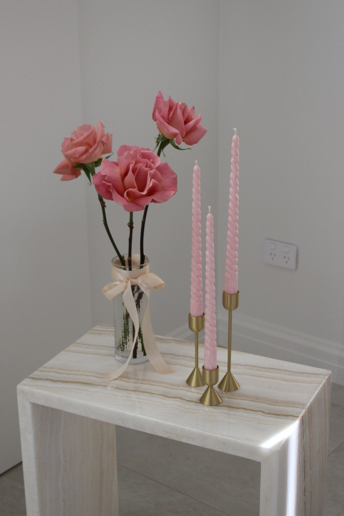 Twisty Ariel taper candle in Peony (pastel pink) colour displayed in a trio on brass candlestick holders set (also known as Lumiere candle holder set). They are positioned on a marble side table near a vase of roses.