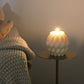Sculptural Brydie candle in Ivory (white) sitting on a brass and marble side table and lit in a dark room. This candle has a smooth braid-like exterior similar to a thick knitted wool blanket.