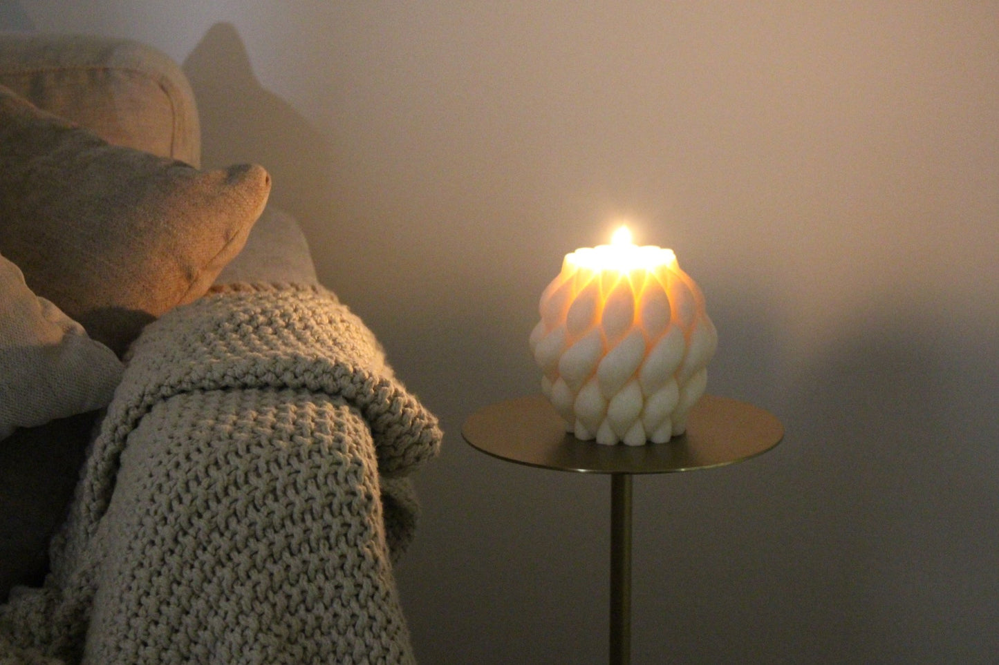 Sculptural Brydie candle in Ivory (white) sitting on a brass and marble side table and lit in a dark room. This candle has a smooth braid-like exterior similar to a thick knitted wool blanket.