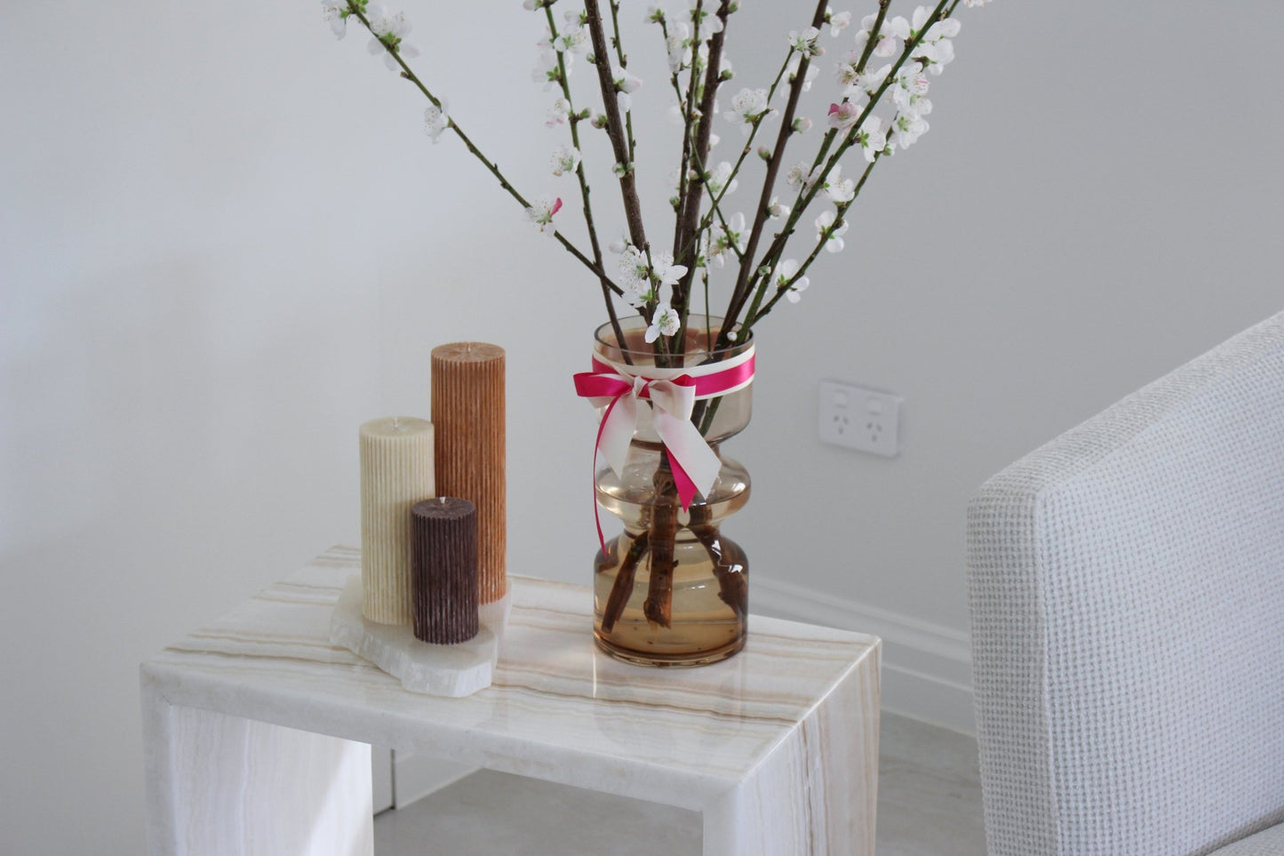 Arrangement of fluted pillar candles shown in white onyx (cream, caramel and cacao (chocolate brown) colours positioned on a side table near a vase of flowers.