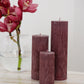 Arrangement of fluted pillar candles shown in Viola (wine) colour positioned on a side table near a vase of flowers.
