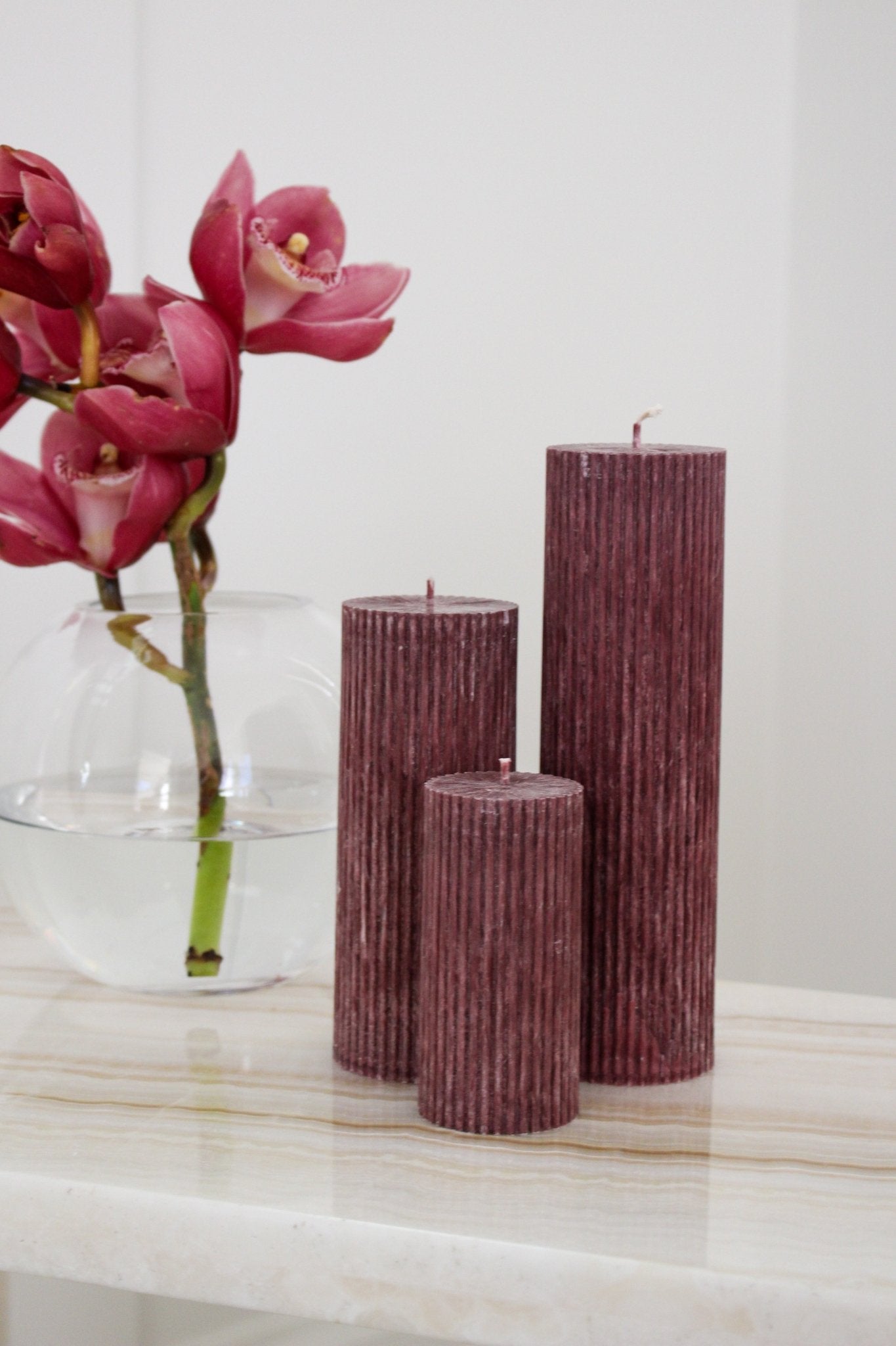 Arrangement of fluted pillar candles shown in Viola (wine) colour positioned on a side table near a vase of flowers.