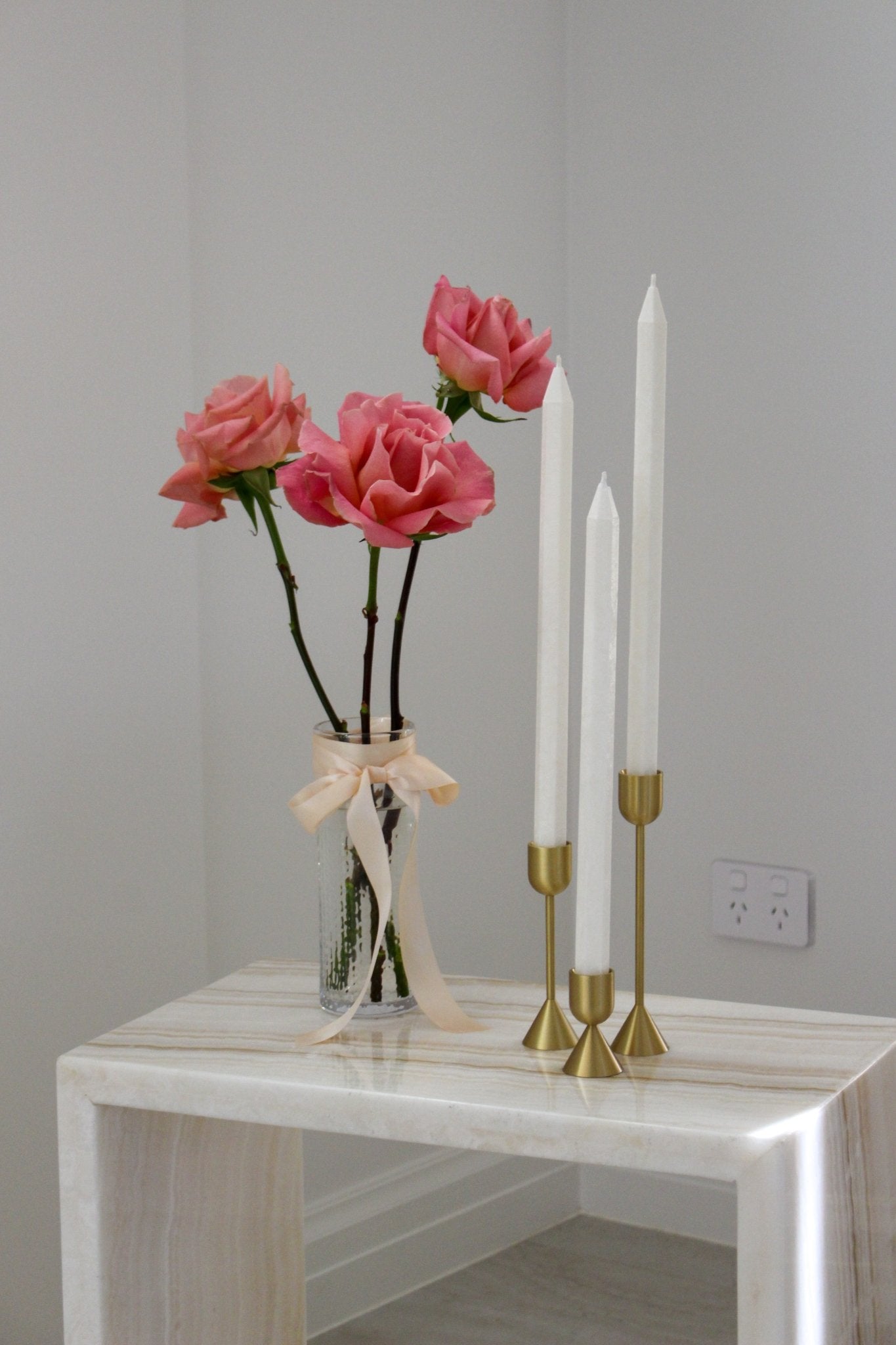 Tall pencil shaped Shiloh taper dinner candle in Ivory (white) colour pictured in a bundle of three. The candles are positioned in brass candlestick holders (known as Lumiere candle holder set) on a marble side table near a vase of roses.